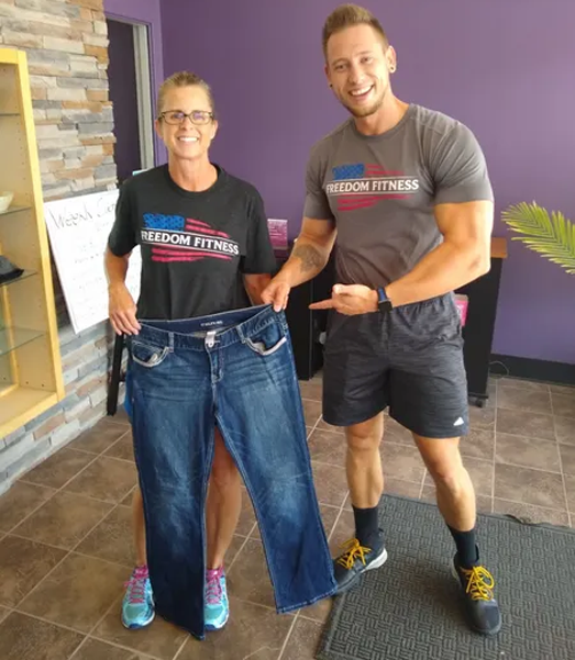 Woman showing weight loss progress holding oversized jeans with trainer at Freedom Fitness in St. Peters, Missouri