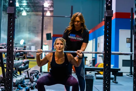 Woman performing barbell squat with trainer spotting at Freedom Fitness gym in St. Peters, Missouri