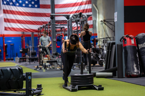 Woman pushing weighted sled on turf during strength training at Freedom Fitness gym in St. Peters, Missouri with American flag in background